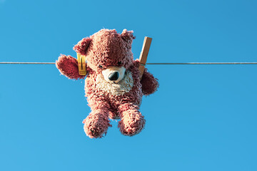 Funny brown teddy bear hanging on a clothesline with wooden clothespin on blue sky background.