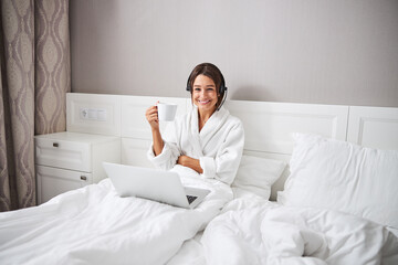 Attractive young Caucasian woman in headphones sitting at the laptop in bedroom