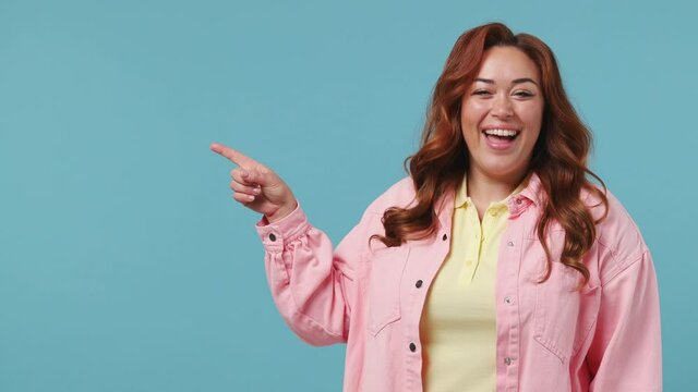 Young beautiful redhead chubby overweight woman 20s years old in pink jacket yellow t-shirt pointing fingers hands aside on copy space mockup promo area isolated on pastel blue color background studio