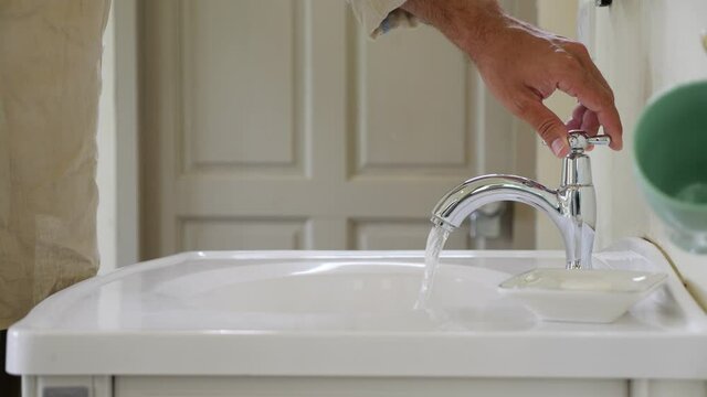Man turn faucet valve by hand and water pour down to bathroom sink. Only one tap with cold water, static camera shows see only male hand, spigot and sink. Blurred door on background