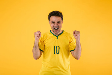 Brazilian soccer fan in a yellow jersey clenches his fists and yells cheering for his team.