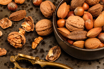 Almonds, walnuts and hazelnuts are sitting in a brown bowl and a nutcracker on a dark brown spotted plate.