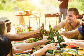 Happy multiracial friends cheering with beers at barbecue picnic meal outdoor - Summer lifestyle, friendship and food concept - Focus on close-up bottles