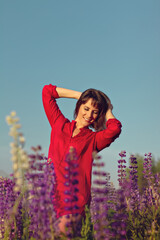 Woman in red shirt with closed eyes in field of lupins and holds glass cup of floral tea in summer