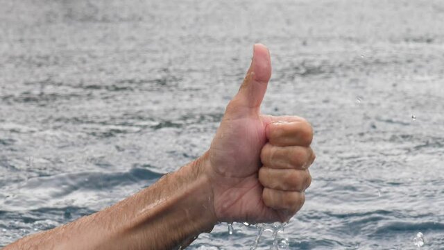 Thumbs up hand jump out from water, close up shot of gesture by man, positive connotation. Closed fist with one finger up, wet and rainy weather, heavy rain pouring down.