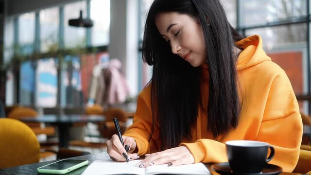 The happy woman in an orange hoodie writing something in a notebook while sitting in a cafe