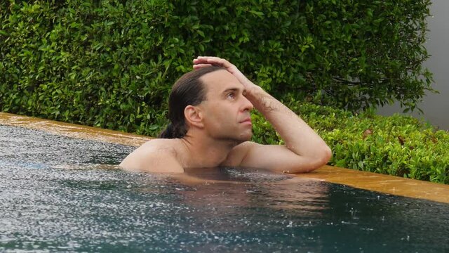 Introvert man stay in swimming pool under heavy rain, stare off into space, move hand down from head. Short slow motion shot of thoughtful freelancer guy, lost in tropical country during wet season