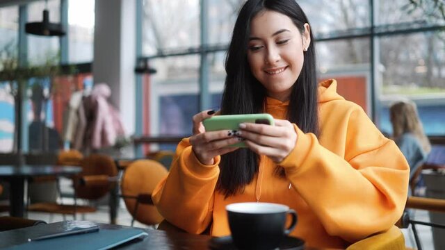 The smiling woman in an orange hoodie playing a game on her phone and rejoicing in victory while sitting in a cafe
