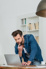 serious teleworker thinking near laptop while standing at workplace