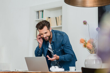 smiling freelancer pointing at laptop while talking on mobile phone