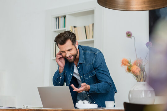 smiling freelancer pointing at laptop while talking on mobile phone