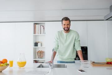 cheerful man smiling at camera near orange juice and sink in kitchen