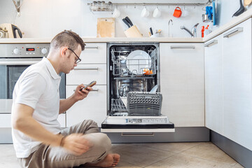 man is studying the instructions in the smartphone for the dishwasher, the husband is doing housework in the kitchen
