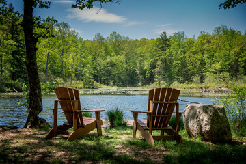 Relaxing inviting scene on summer day with two chairs facing a pond with forest in background and...