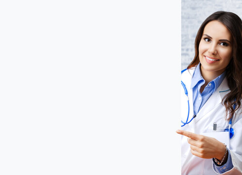 Portrait image of happy smiling female doctor pointing blank signboard with copy space, over white brick loft wall, at office. Young brunette woman in medical, clinic, healthcare concept.