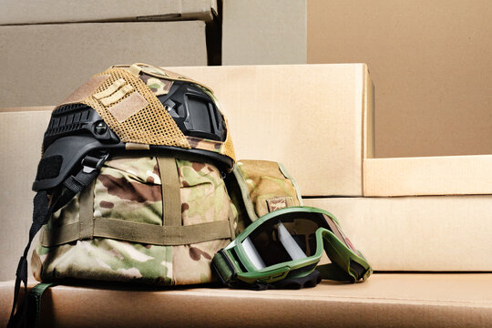 Photo of various military camouflaged armored helmets with covers and soldier googgles. laying on stacked cardboard boxes.