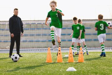 Obraz premium Young sports boys in green sportswear running and jumping over training obstacles on pitch. Soccer youth team playing football training in the summer. Sports activities for kids. Coach with school boy