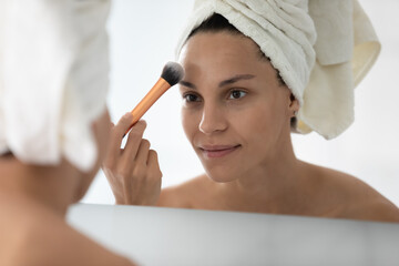 Focused positive girl with head wrapped in towel putting makeup on smooth silky facial skin after shower, using cosmetic brush with powder. Young woman applying foundation on face. Mirror reflection