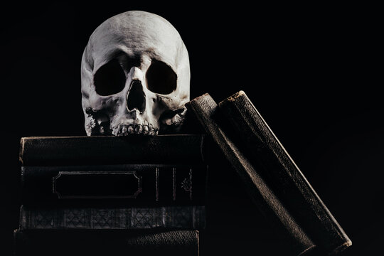 Photo of witchcraft human skull laying on old books and stacked books on black background.