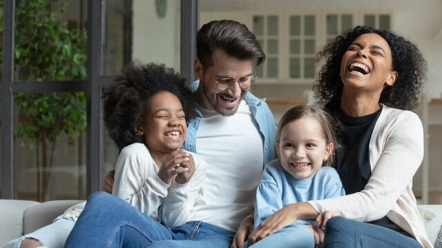 Overjoyed young multiracial family with two small daughters relax on sofa at home laughing joking. Happy multiethnic mom and dad rest together with small diverse children. Adoption concept.
