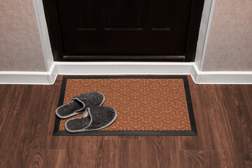 Blank doormat before the dark door in the hall. Home slippers on the rug. Top view of mat on wooden...