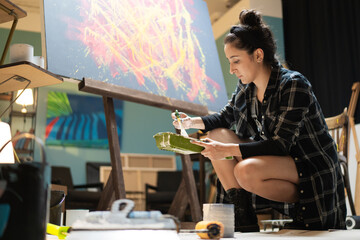 A artist is working in her studio, school, and art class on a new piece of painting related to abstractionism, modernity. The girl crouches on the floor picks up a tray scooping paint onto a brush.