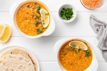 Masoor dal. Indian spicy red lentil soup in two white plates, roti (chapati) half a lemon, sliced green coriander, gray napkin and dry raw lentil are on white wooden table.