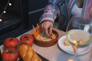 Person eating hummus with carrots on a folding table next to a van parked outside. - obrazy, fototapety, plakaty