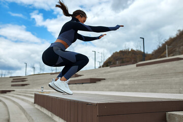Fototapeta na wymiar A young woman runner-athlete in a dark tight-fitting suit runs down the city stairs, jumps up the steps. city workout