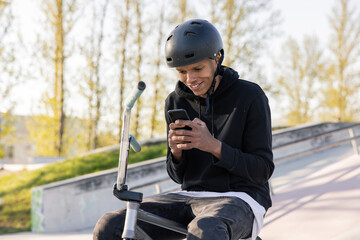 Guy sits in helmet with bmx bike in park on ramp, at skatepark with phone in hand and texts friends he hangs out with to do tricks