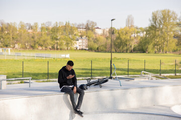 Boy sits in helmet with bmx bike in park on ramp, at skatepark with phone in hand and texts friends...