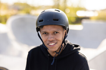 A young wearing a helmet stares into the camera with dark brown eyes with white, straight teeth on top. In the background you can see the ramp and the greenery of the park surrounding the ramp