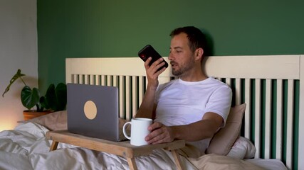 Man sitting in bed with laptop, speaking voice mails on smartphone - Powered by Adobe