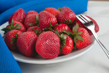 A white plate on a table, full of strawberries with a fork on top and a  blue tablecloth on th side. Side view