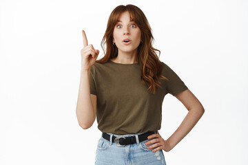 Excited young woman suggesting plan, raising finger pointing up, gasping and looking surprised, astonished by advertisement upwards, standing against white background