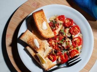 A cherry tomato snack on a white plate with toasted bread with butter and a fork on the side over a wooden cutting board and a glass cup of olive oil, a spoon and a blue napkin. Olive oil dressing