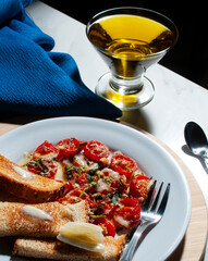 A cherry tomato snack on a white plate with toasted bread with butter and a fork on the side over a wooden cutting board and a glass cup of olive oil, a spoon and a blue napkin. Olive oil dressing
