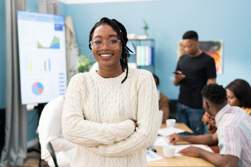 Portrait of a smiling dark-skinned girl wearing a warm sweater with her arms crossed over chest, woman wears glasses and gold earrings, her hair is braided, a business meeting in office in background - obrazy, fototapety, plakaty