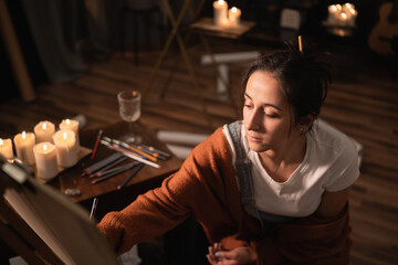 Female sits in the middle of the room amidst scattered failed drawings and candles. The beautiful girl is focused on making the perfect painting, a pencil sketch.