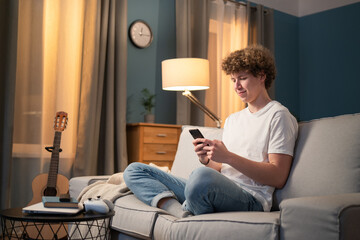 Young boy with curly dark hair sits on couch in living room in evening and sends text message to girlfriend, friends, acquaintances, posts on social media