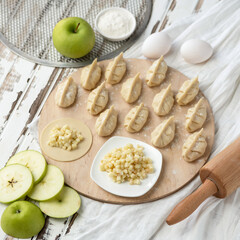 Cooking process. Sweet Dumplings with green apple filling on Raw dough circles at wooden cutting board. Ingredients and rolling pin on rustic background. Convenience Food concept. Flat lay.