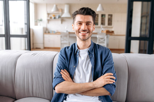 Portrait of attractive peaceful joyful young caucasian man sitting at home with arms crossed on living room sofa, wearing casual stylish clothes, looking at camera and smiling friendly