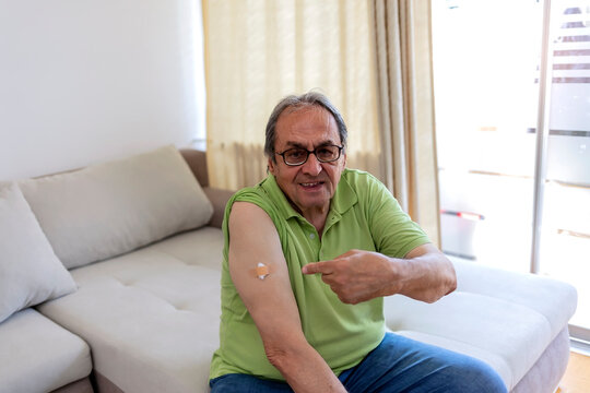 Portrait of a proud cheerful senior man who had just been vaccinated sitting on sofa at home. Senior man showing vaccinated arm. Coronavirus vaccination concept of man received the COVID-19 vaccine.