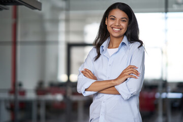 Portrait of a young happy attractive business woman or female office worker keeping arms crossed, looking at camera and smiling while working in the modern office