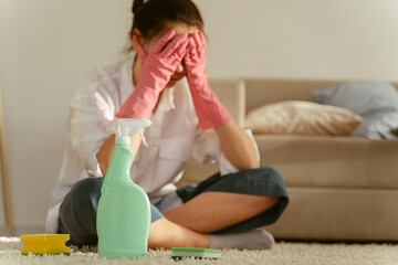 Tired young woman desperate to wash stains on carpet at home, sitting alone on floor covering face with her hands or crying, focus on cleaning supplies. Housewife refusing to do chores