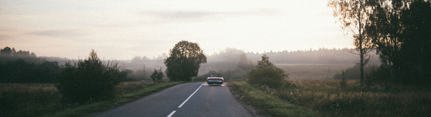 A retro car drives along the road at dawn. The convertible drives off into the distance on a...