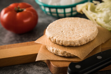 Vegan Vegetarischer Burger Patty roh auf Holz Schneidebrett und Messer mit Zutaten Eisbergsalat und Tomate auf dunkel Beton Hintergrund
