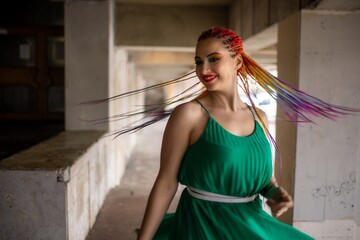 A girl with a happy smile in a spring dress with bright makeup and multi-colored braids is spinning while walking along the warm streets of the city