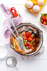 Dutch baby pancake with fresh strawberry berry and sprinkled with icing sugar powder in red pan on white kitchen background. Top view