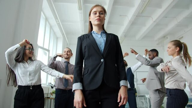 Low angle shot of ambitious woman standing in office with serious face while coworkers dancing around enjoying party. Emotions and celebrations concept.